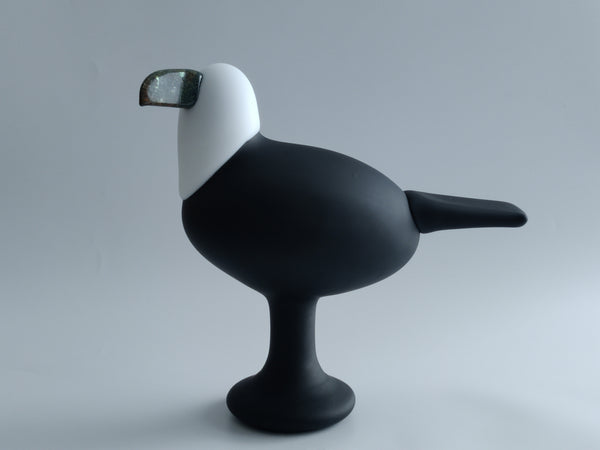 A modern bird-shaped object with a matte black body and leg, and a white head, featuring a dark, textured beak, set against a plain light gray background.