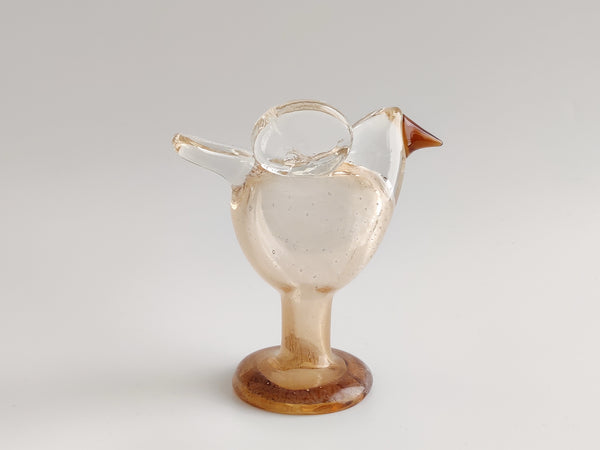A small bird-shaped glass vessel with a transparent body, amber-brown base, and brown beak, photographed on a light gray studio background.