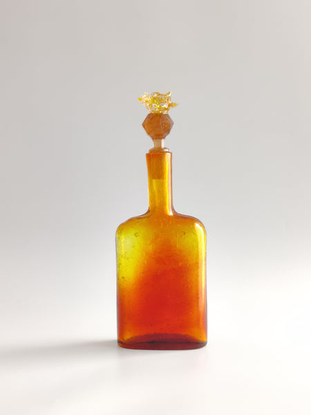 A small amber glass bottle with a narrow neck, featuring a faceted orange stopper topped with a gold-toned decorative element, photographed against a light gray studio background.