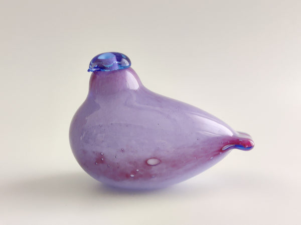 A small hand-blown glass bird figurine in lavender purple with a blue beak and a pinkish-red tail, set against a plain light gray background.