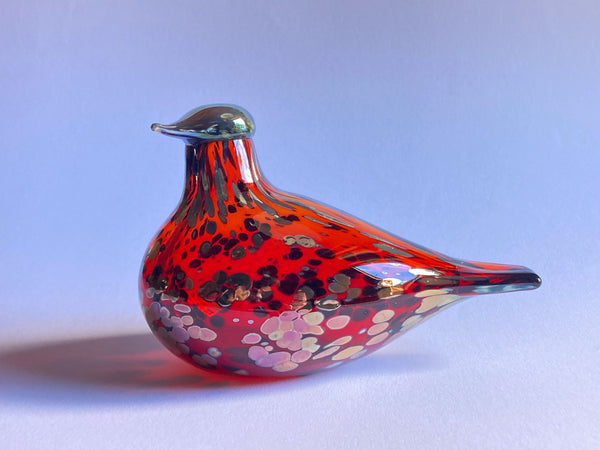 A hand-blown glass bird figurine with a vibrant red body, speckled with black, white, and pink, and finished with a metallic silver beak, set against a soft blue gradient background.