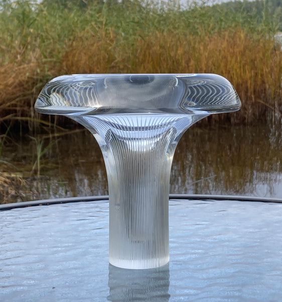 Clear art glass object with ribbed texture on a reflective surface with grass and water in the background
